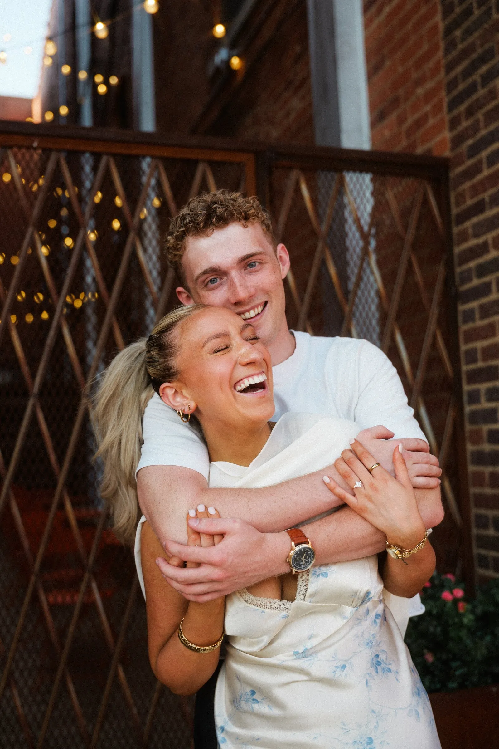 A couple laughing, one hugging the other from behind, in a brick courtyard
