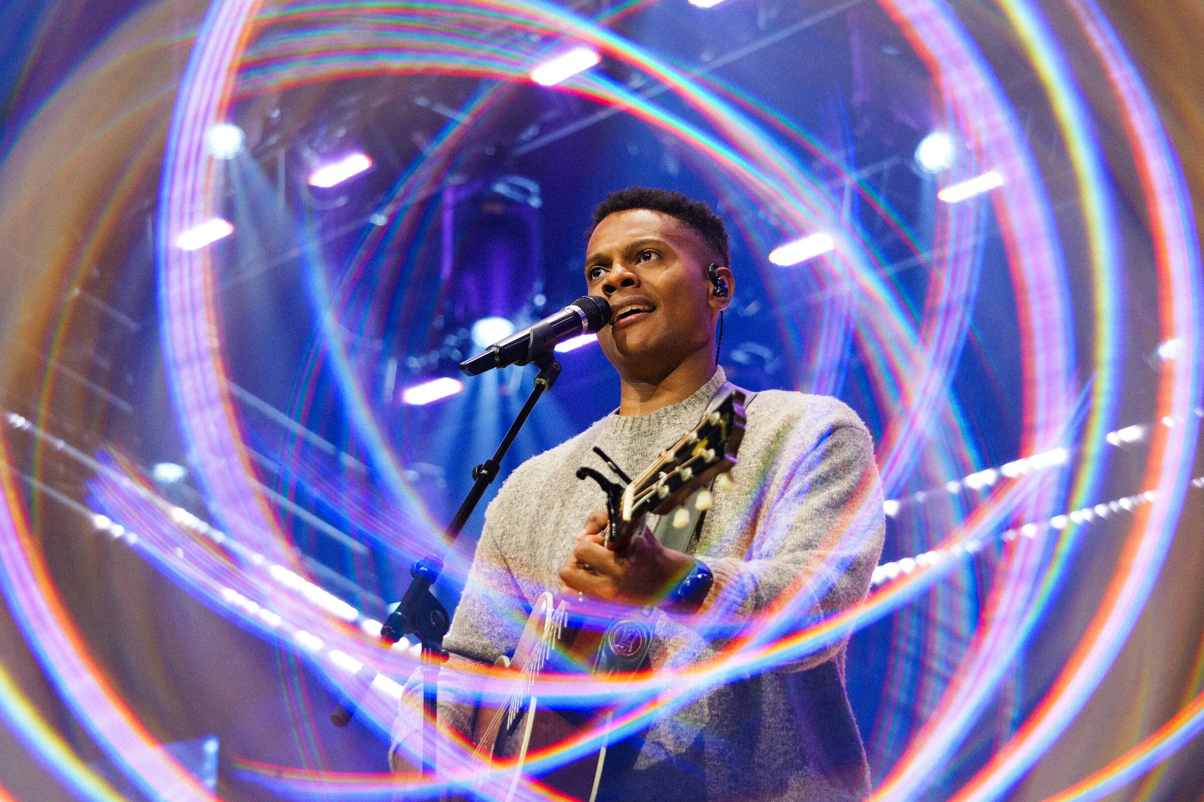 A guitarist singing at a mic, wrapped in rings of stage light