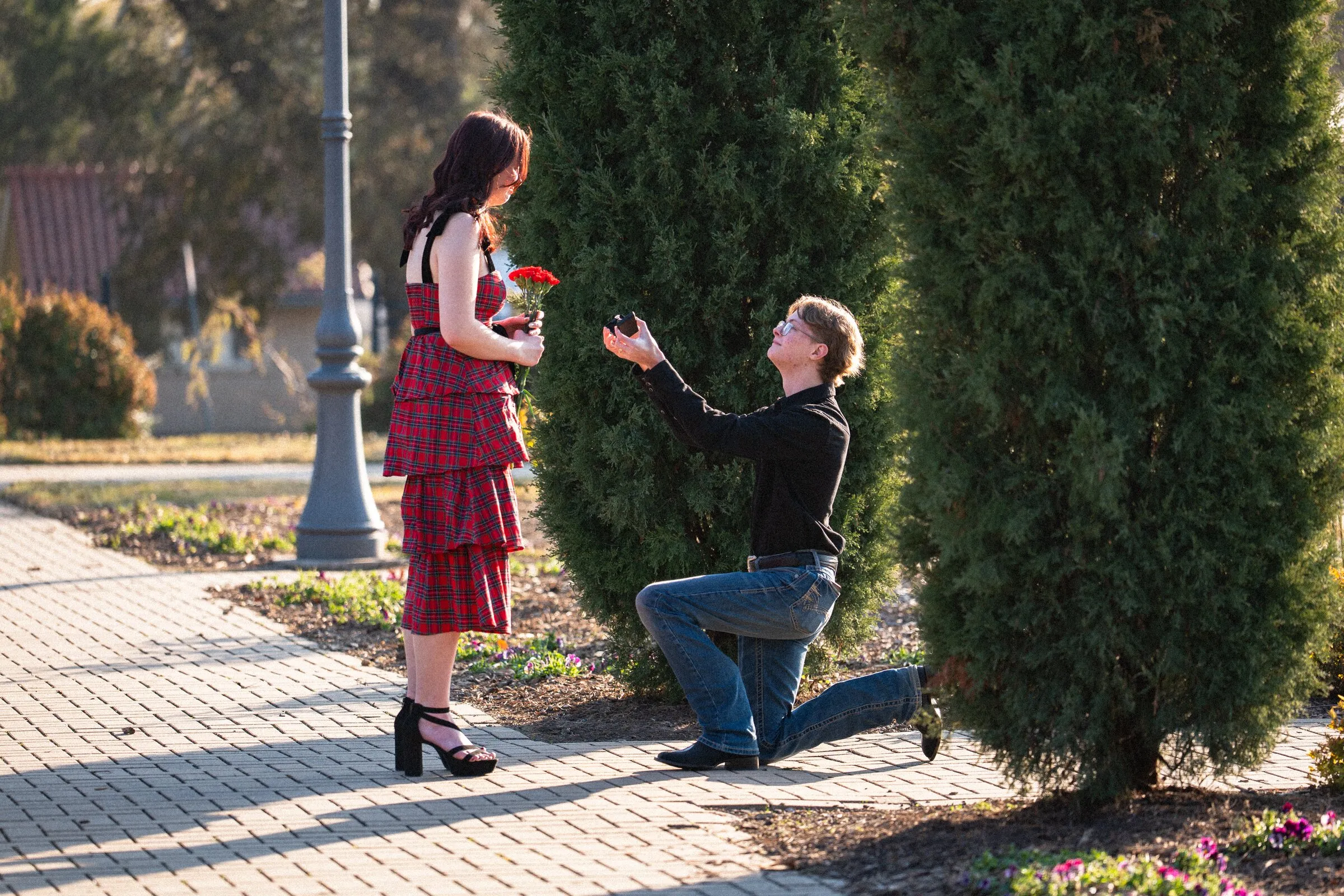 A man on one knee proposing to a woman in a red dress on a brick sidewalk