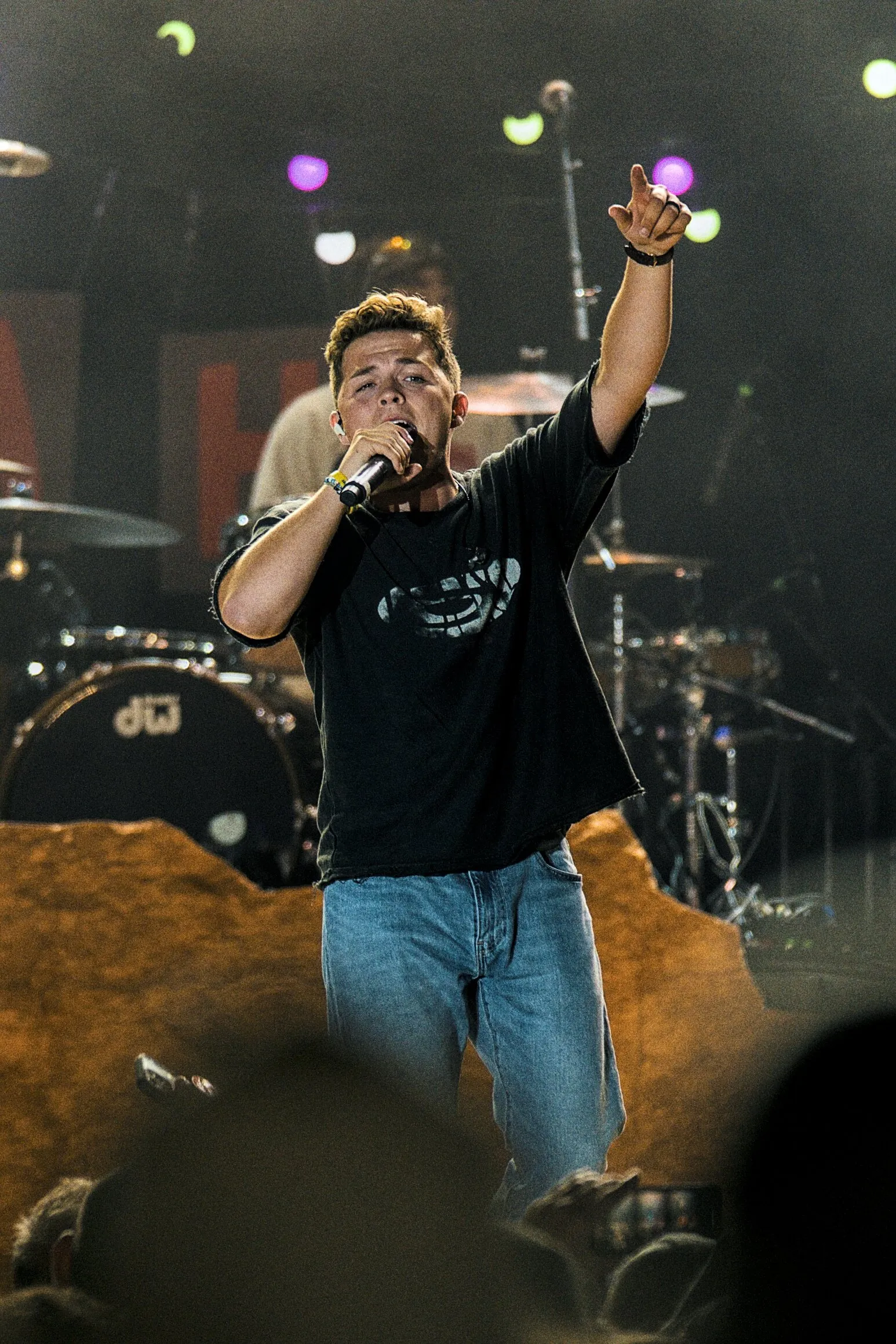 A performer on stage, one arm raised to the crowd, drum kit behind