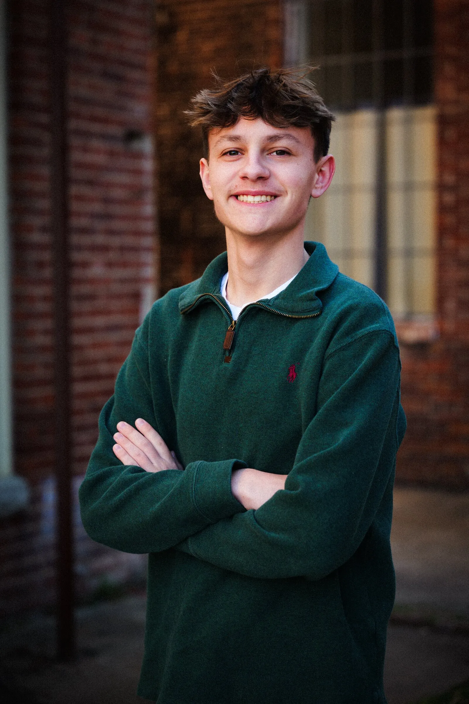 A senior in a green quarter-zip, arms crossed, smiling in front of a brick building