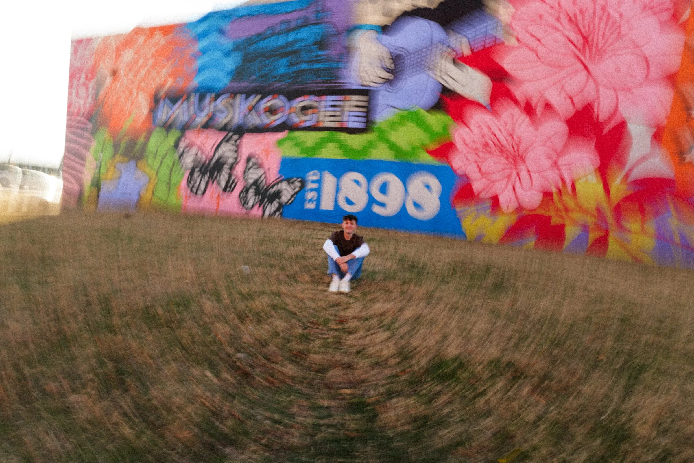 A senior sitting in front of a bright Muskogee mural, shot from low and wide