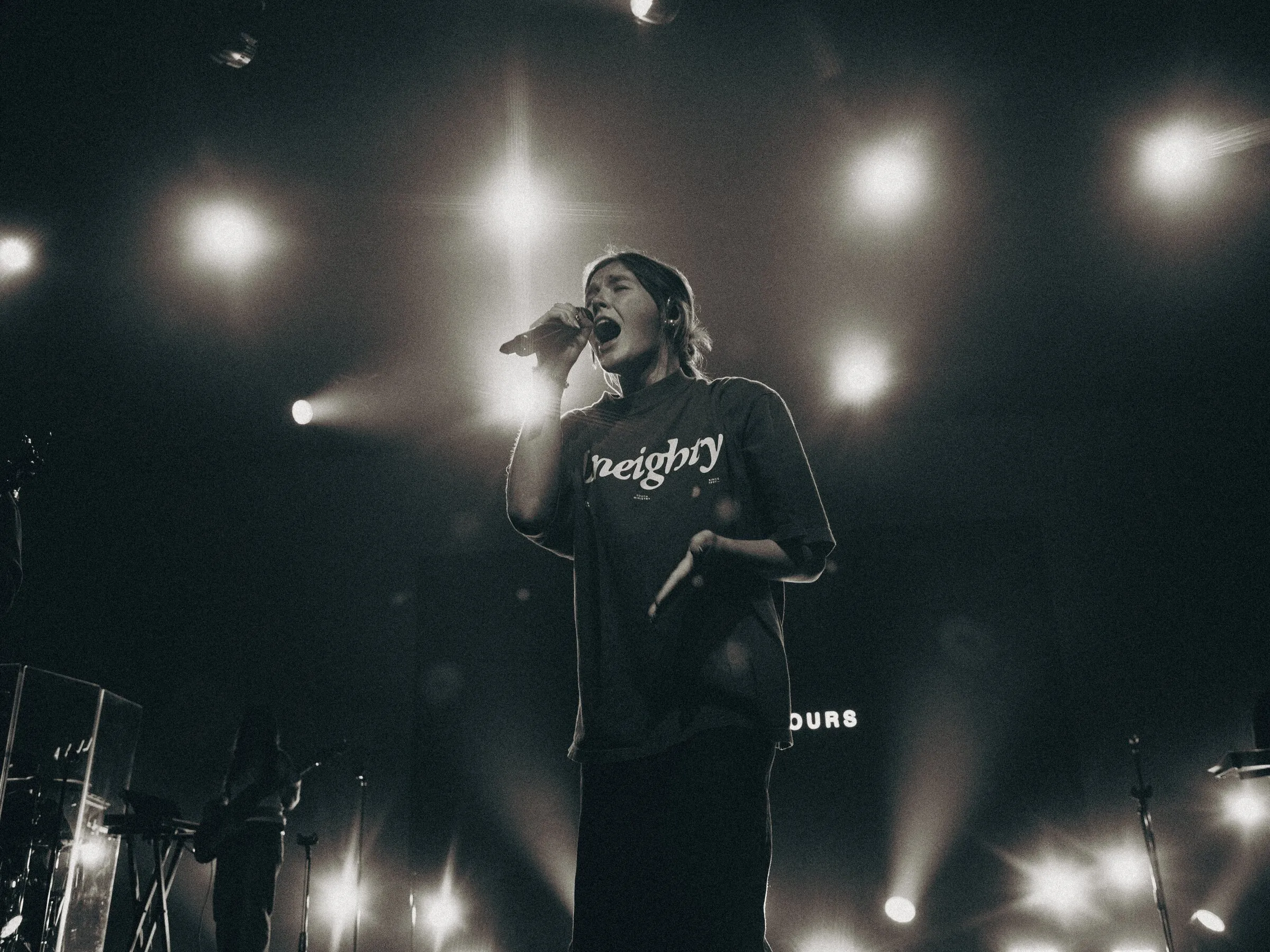 A singer at the mic under a grid of stage lights, black and white
