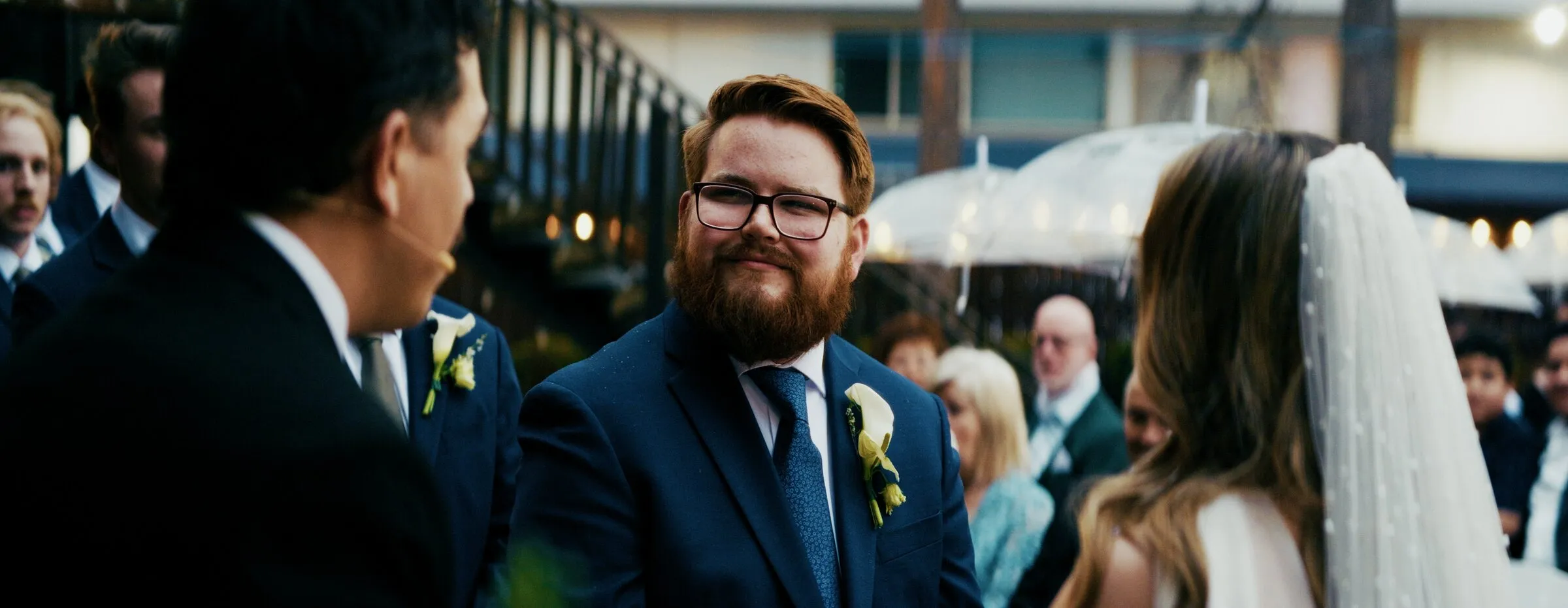 A groom in navy at the altar, grinning at the bride as guests in the rain look on under umbrellas