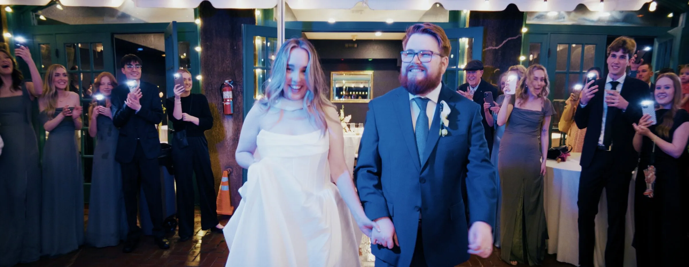 Bride and groom walking into the reception hand in hand as guests cheer and raise their phones