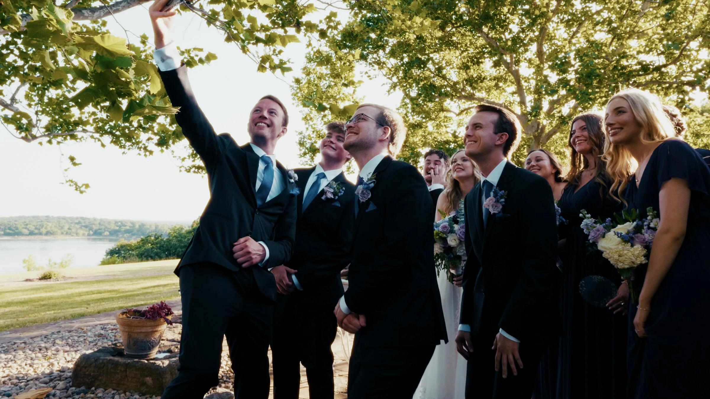 The wedding party laughing together under a big tree by the lake