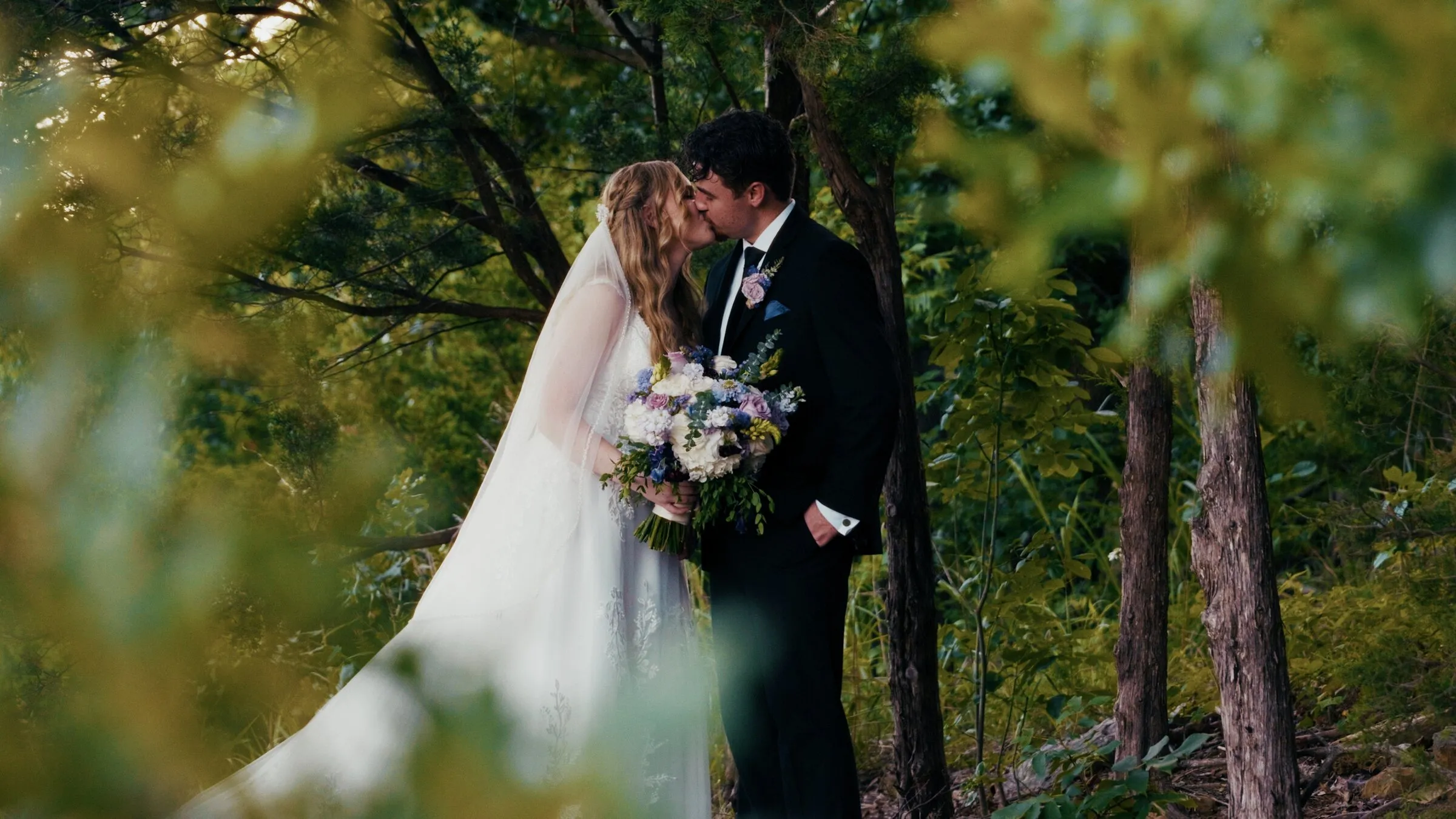 Bride and groom kissing under the trees, seen through green leaves
