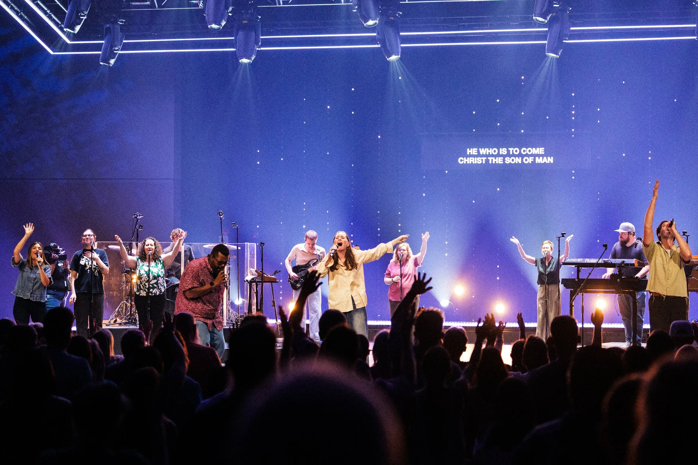 A worship band on a wide stage under blue light, arms raised, crowd in the foreground